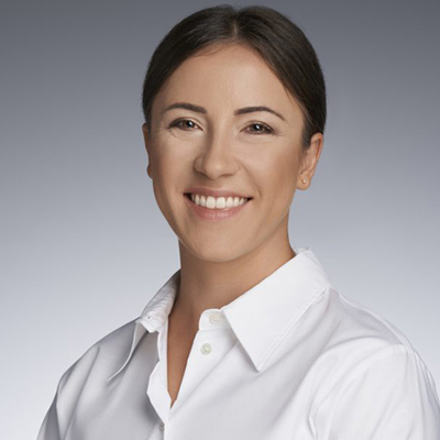 Professional headshot of a woman with dark hair pulled back, wearing a white collared shirt, smiling warmly at the camera against a neutral gray background.