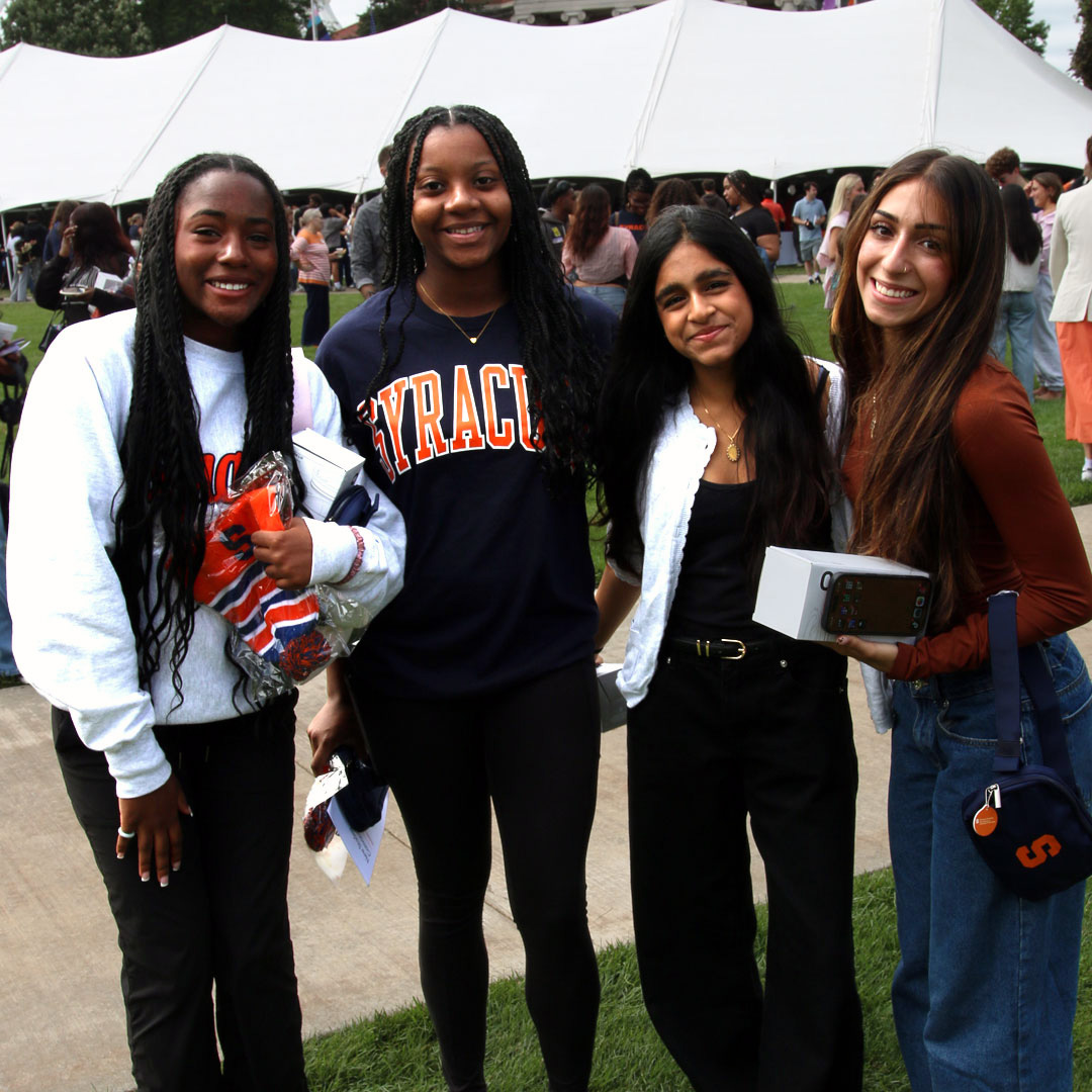 Four people standing outdoors in front of a white tent, two wearing Syracuse apparel, with others in the background under a white tent at a campus event.