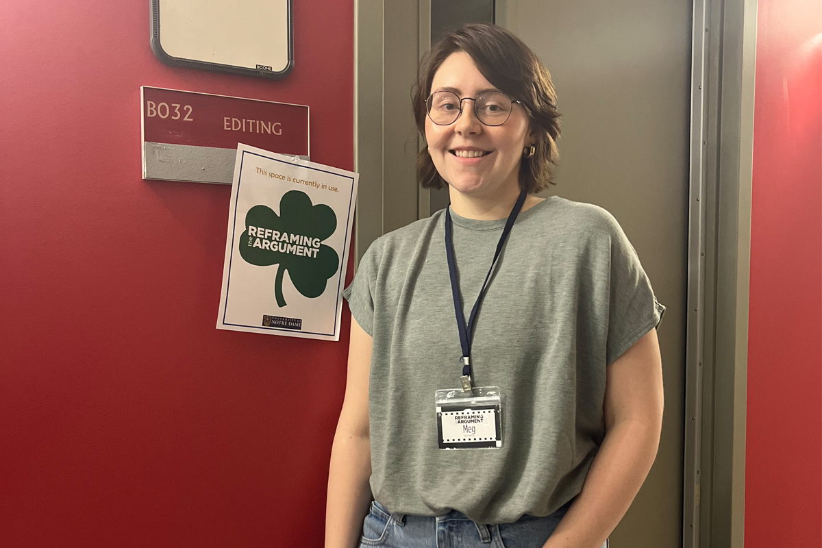 A person with short brown hair and glasses stands smiling in front of a red door marked "B032 EDITING." They're wearing a gray t-shirt and jeans with a name tag lanyard around their neck.