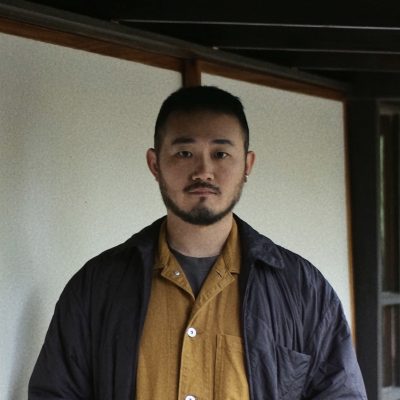 Man with short dark hair and facial hair wearing a mustard yellow shirt under a dark jacket, standing in an interior space with exposed wooden beams and white walls.