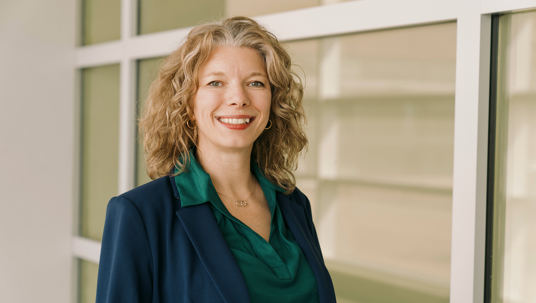 Professional portrait of a woman with curly blonde hair wearing a navy blazer over a green blouse, smiling at the camera against a modern office building background with large windows.