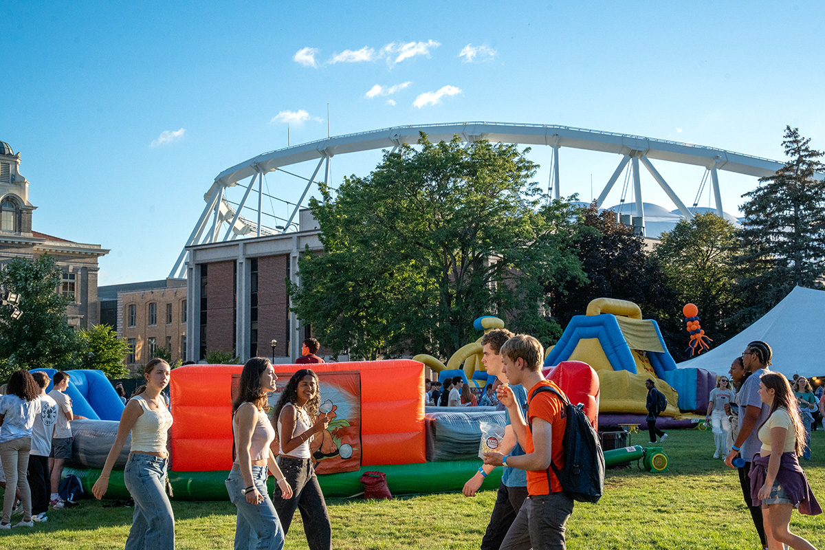 Group of people outdoors on a sunny day near inflatable structures, with a large white arch, trees, and buildings in the background.