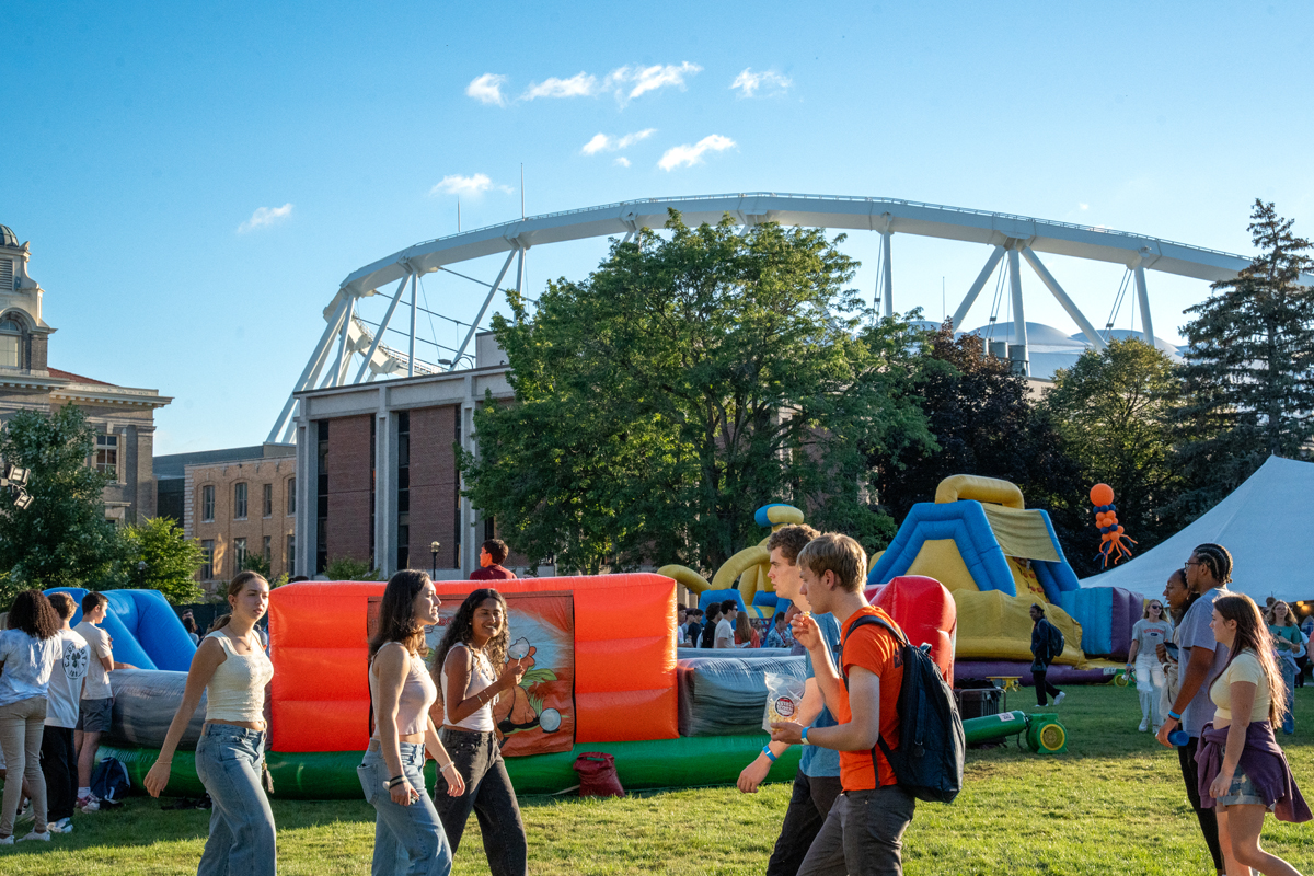 Students enjoy a campus event on a sunny day, with colorful inflatable attractions set up on a grassy field near brick buildings and a large white stadium arch.