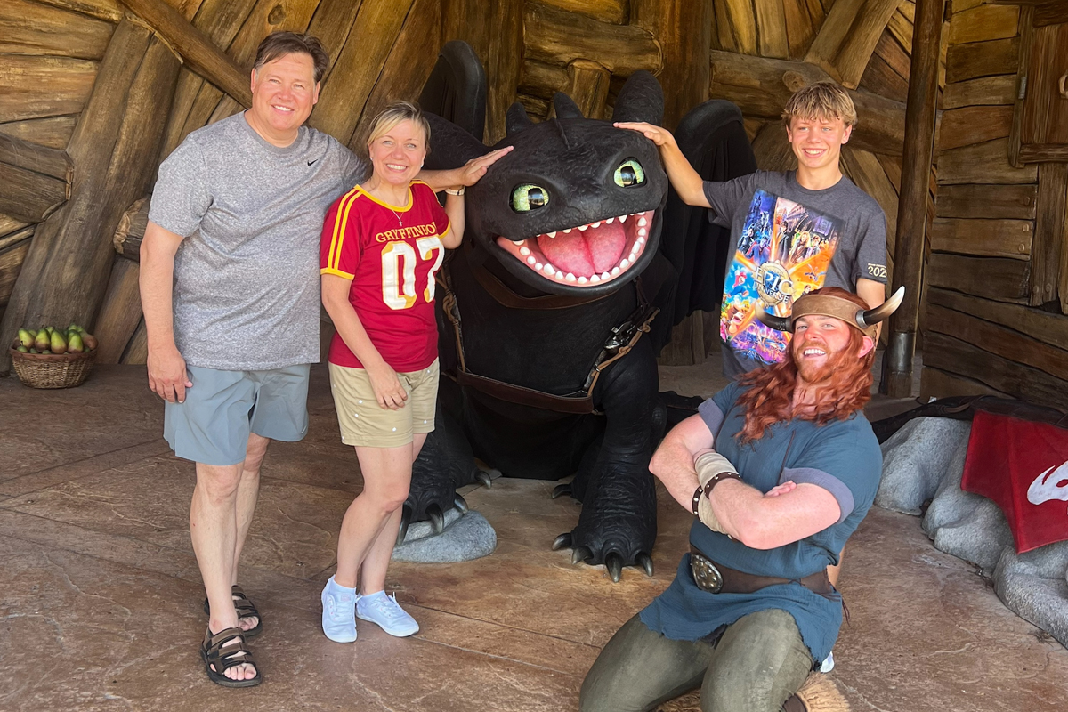 Family of four posing with large black dragon character (Toothless from How to Train Your Dragon) and Viking character actor inside rustic wooden building at theme park.