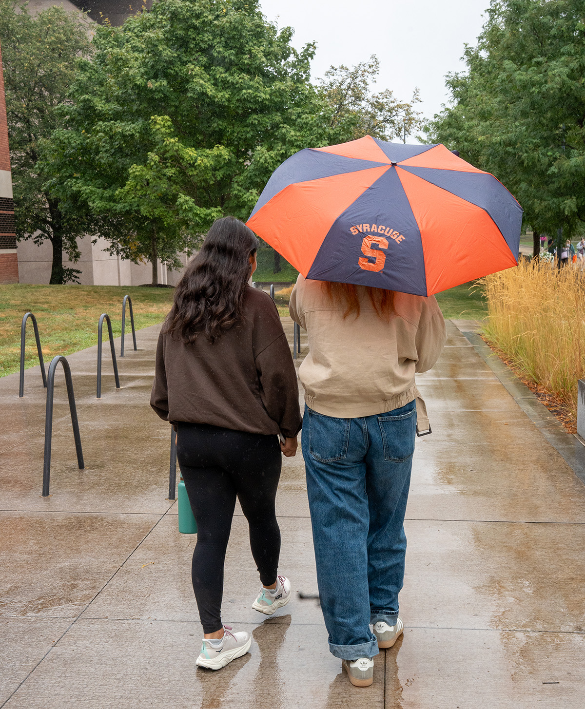 Two people walking on a wet sidewalk under a Syracuse umbrella, with bike racks and tall grass nearby.