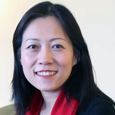 A professional headshot of a person with shoulder-length black hair, smiling warmly at the camera. She's wearing a red top or blouse.