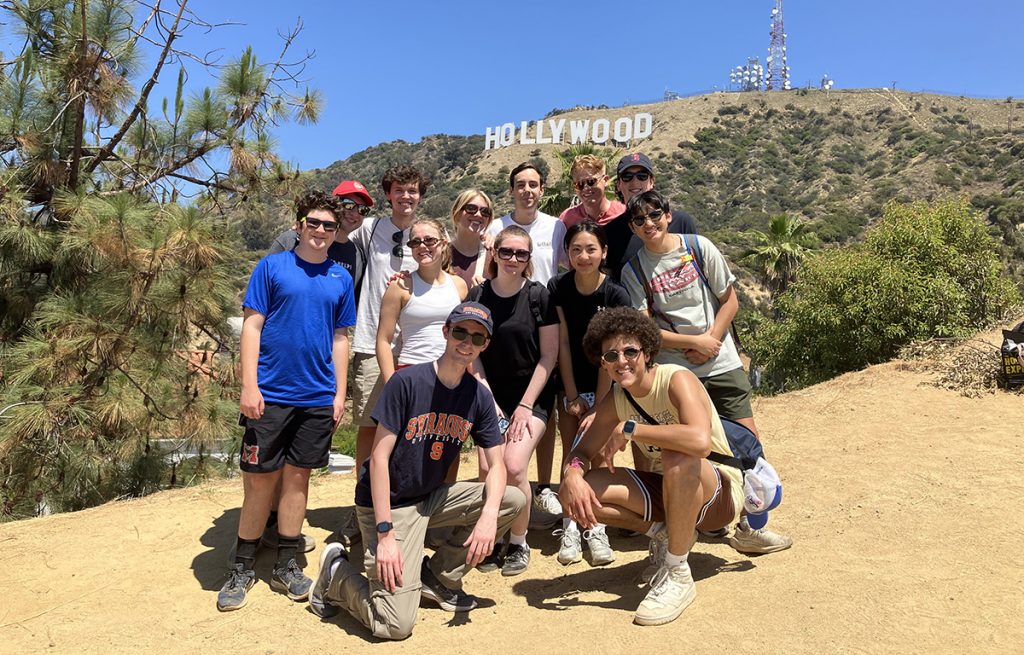 Group of 13 people posing on a dirt path with the Hollywood sign in the background.