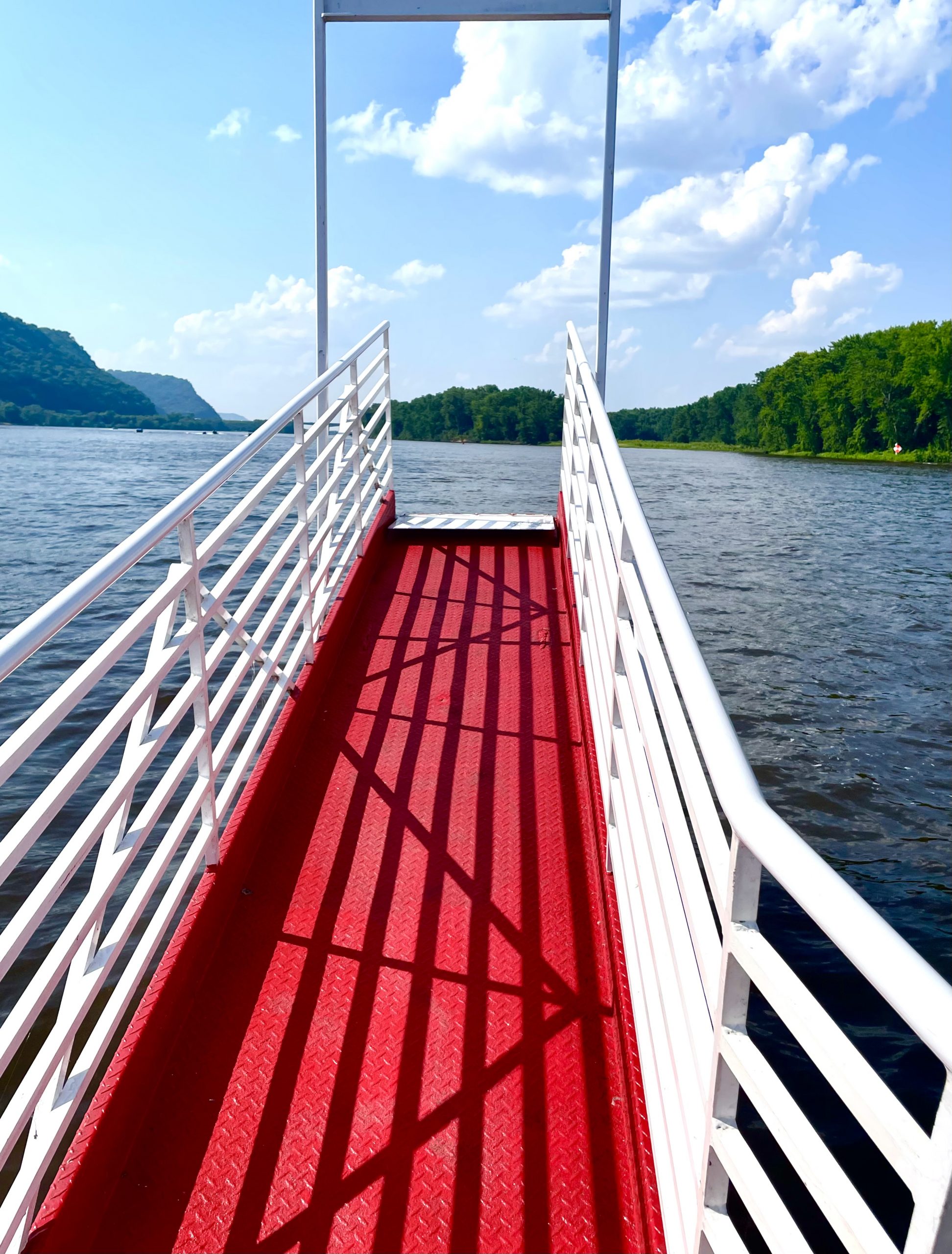 View from boat deck with bright red non-slip flooring and white railings, looking out over calm lake surrounded by forested hills under blue sky with white clouds.