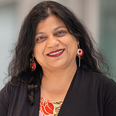 Professional portrait of a woman with long dark wavy hair, wearing red statement earrings, a black blazer over a red and white patterned top, smiling warmly at the camera with a blurred office background.
