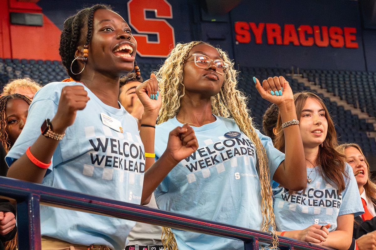 Welcome Week Leaders in light blue shirts cheering in a stadium with 'SYRACUSE' displayed on a dark blue wall behind them.