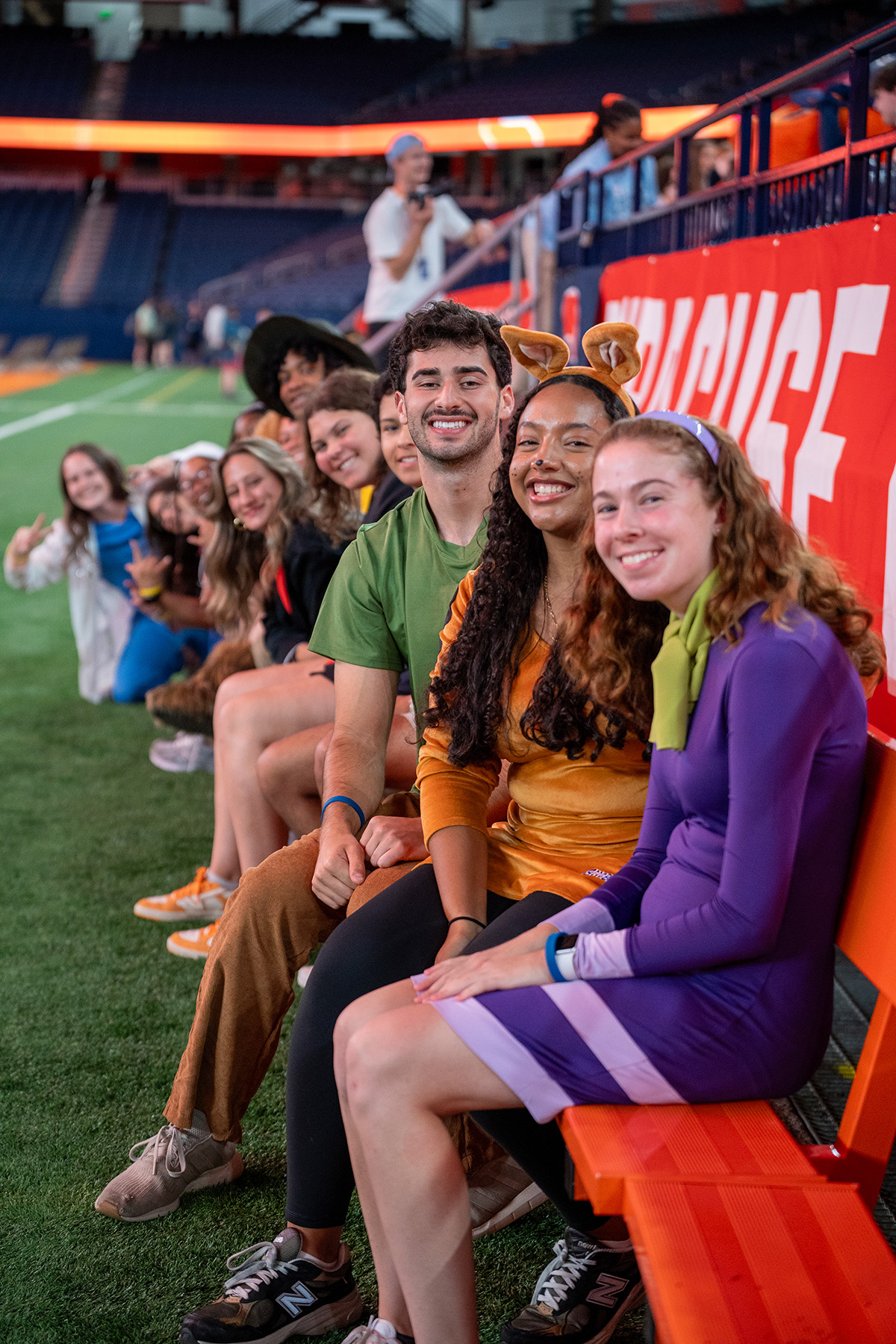Group of people in costumes seated on a bench inside a sports stadium, with field, stands, and spectators in the background.