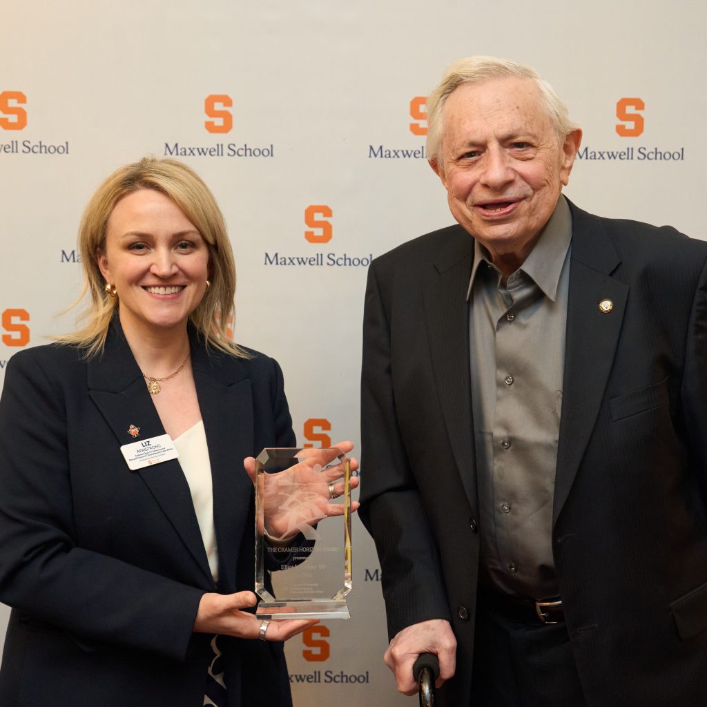 A woman in a dark business suit holds a clear glass award while standing next to an older man in a dark suit jacket and light shirt. They are both smiling at a formal event, positioned in front of a backdrop featuring repeated "Maxwell School" logos with orange "S" letters.