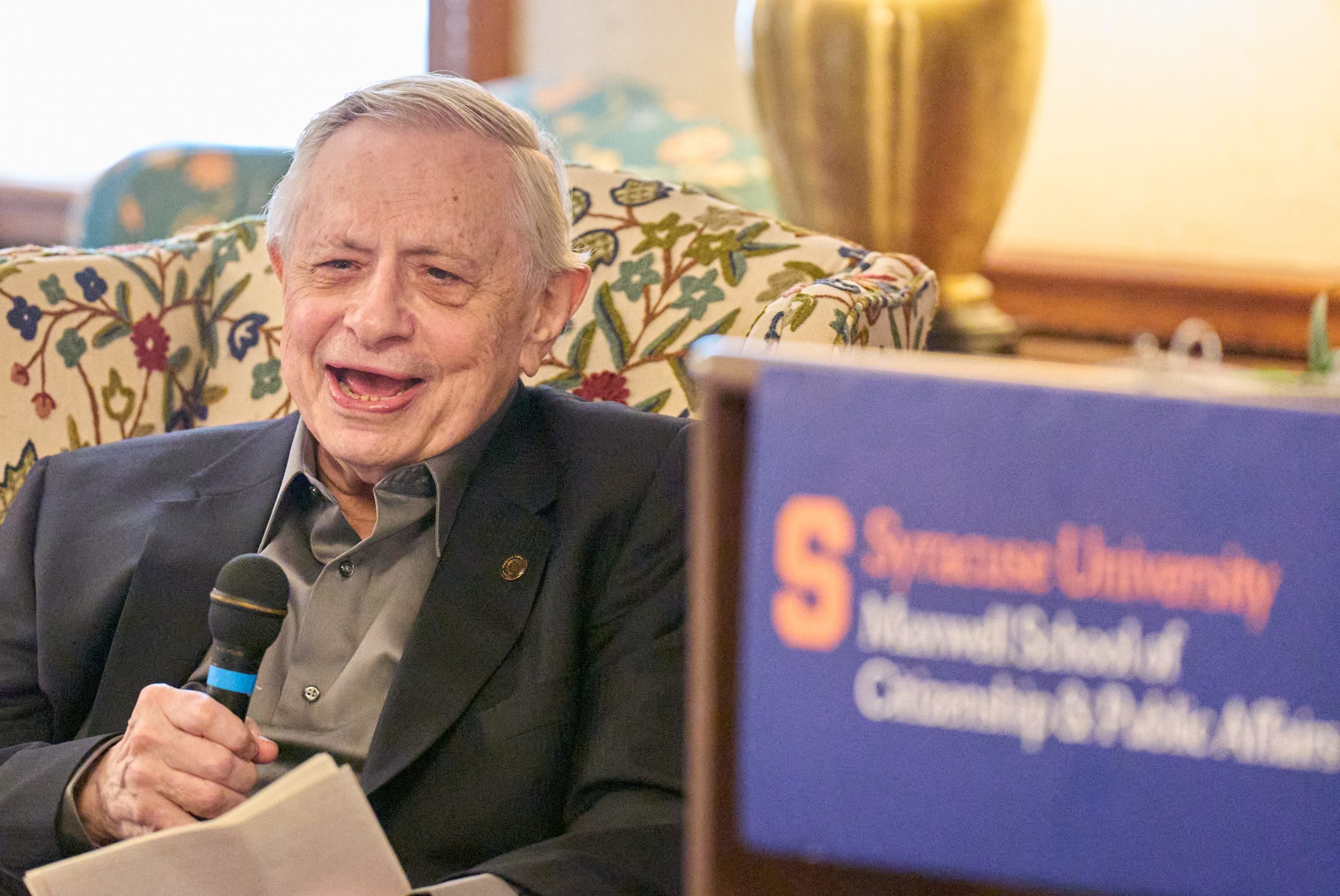 A man in a dark suit jacket and olive shirt sits on a floral-patterned chair, holding a microphone and speaking at a Syracuse University event.