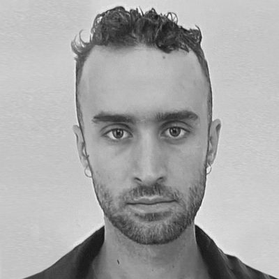 Black and white headshot of a man with curly hair on top, shaved sides, facial hair, and earrings, wearing a dark collared shirt.