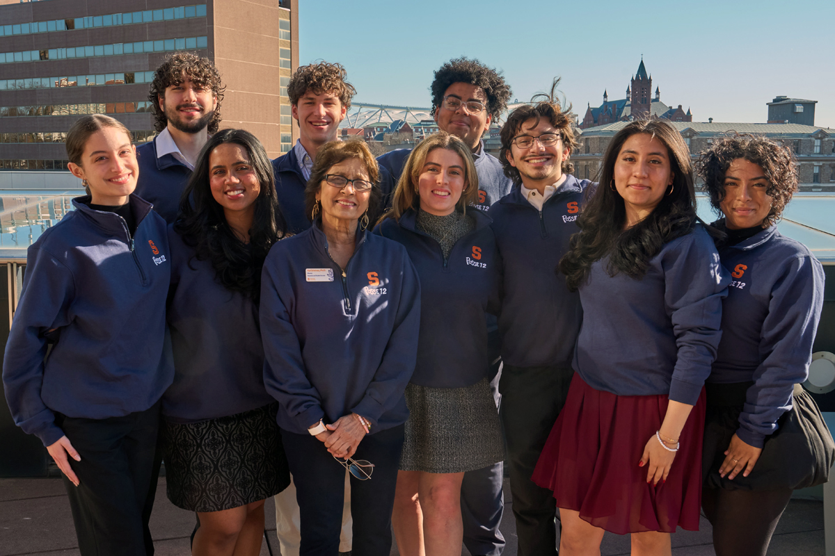 A group of 10 people wearing navy blue jackets with Syracuse University logos pose together on an outdoor terrace or rooftop, with urban buildings and a clear sky visible in the background.