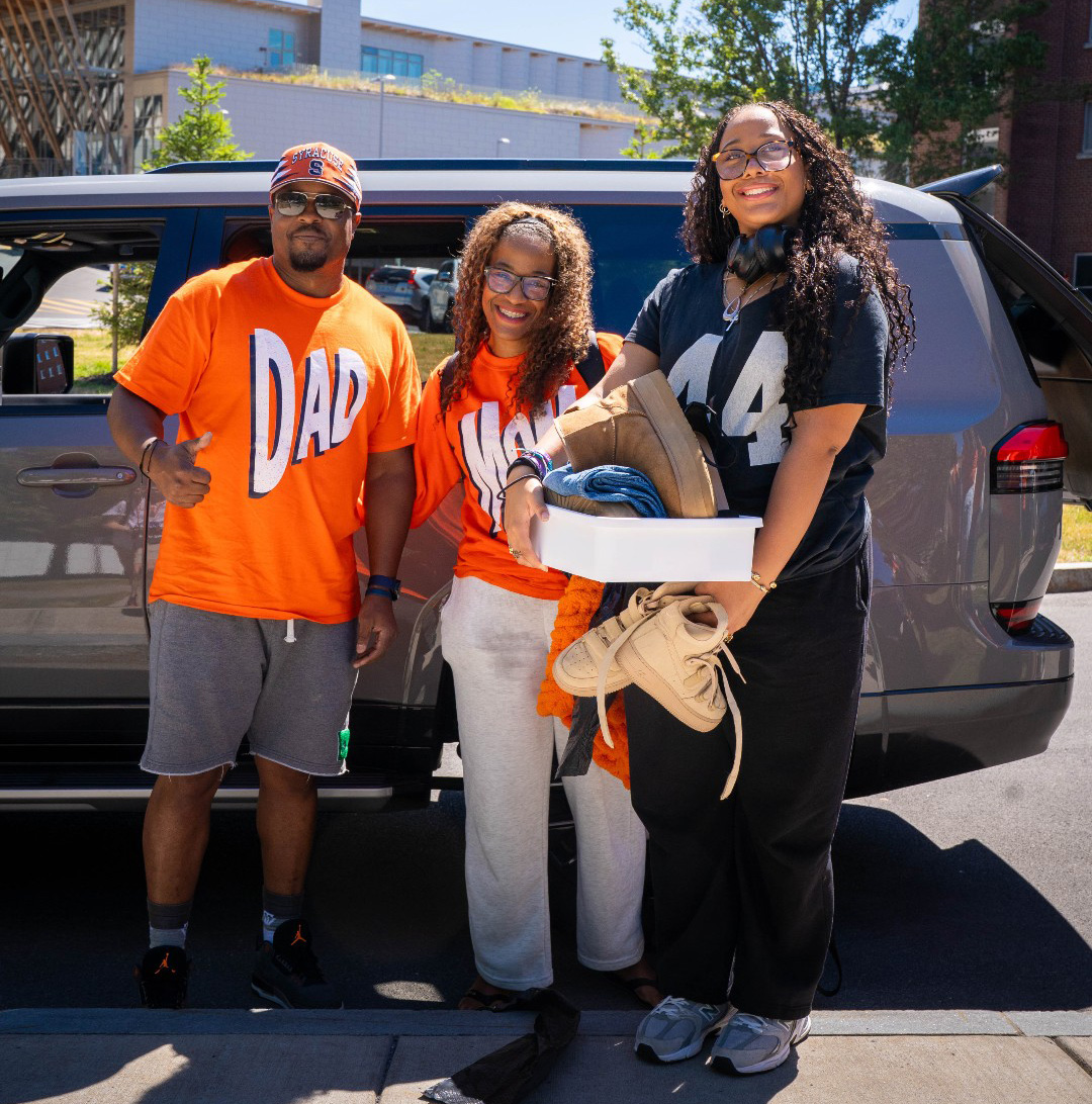 Three people standing in front of an SUV, wearing Syracuse-themed shirts labeled 'DAD,' 'MOM,' and '44,' with one holding shoes and a box.