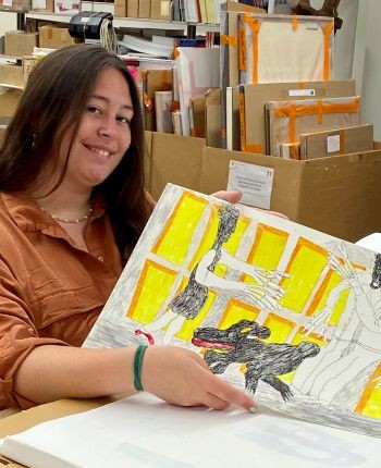 Samantha Hefti G ’25, a museum studies graduate, holds a colorful artwork in a storage room at the University's Special Collections Research Center where she is surrounded by boxes and art supplies.