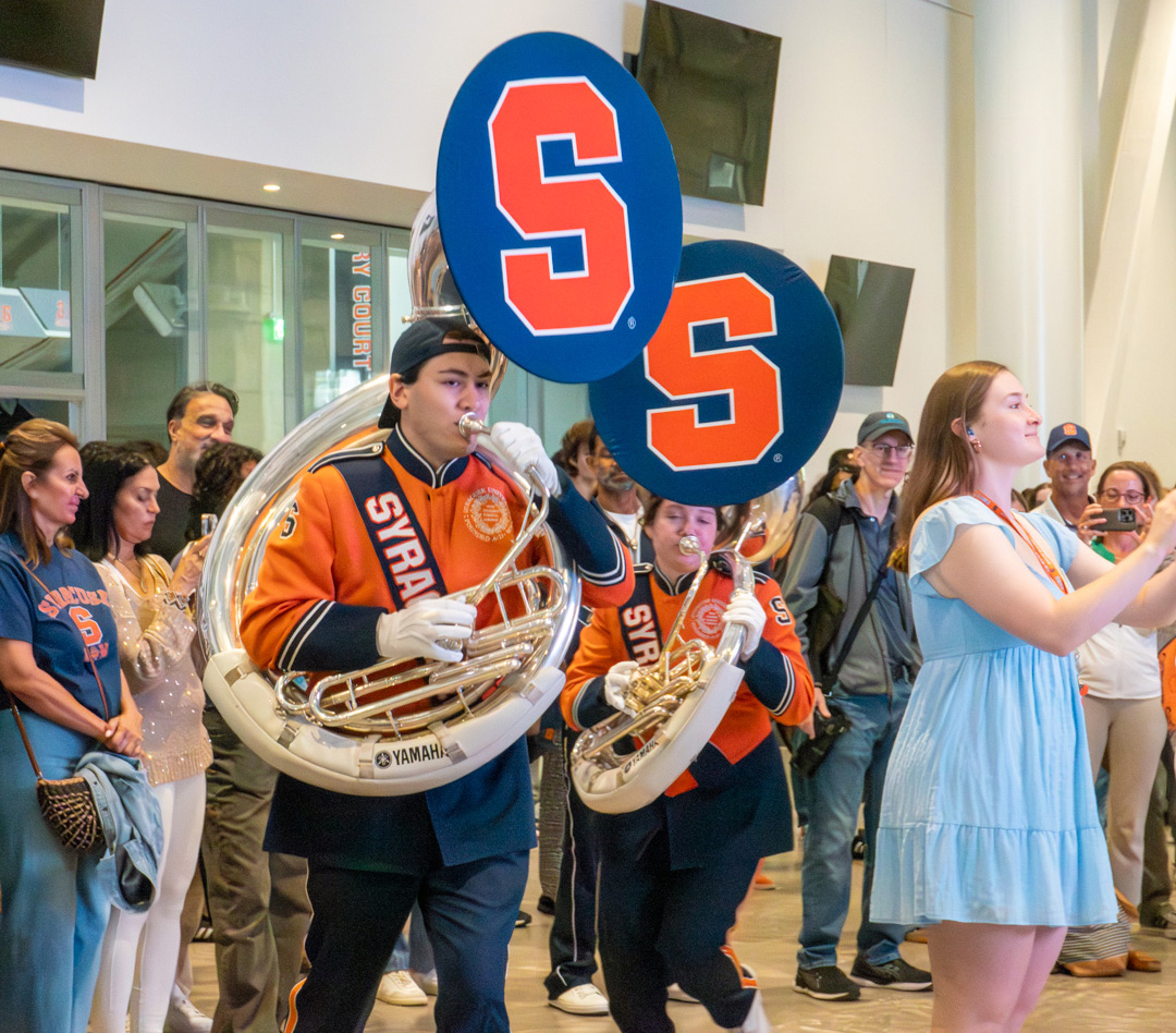 Syracuse marching band performing indoors in orange and blue uniforms, with sousaphones and a cheering crowd.