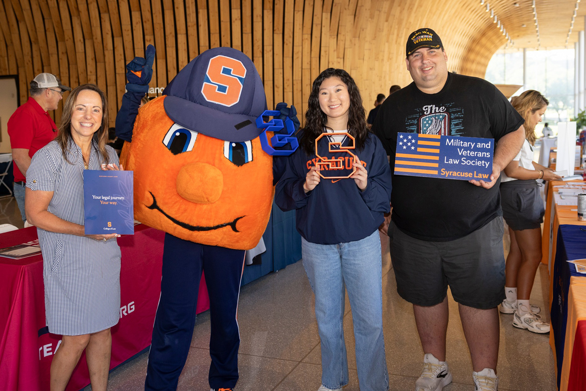 Three individuals pose at a Syracuse University Law event with Otto including a woman with a law booklet, another with a university pennant, and a man holding a Military and Veterans Law Society sign, all set in a modern indoor venue.