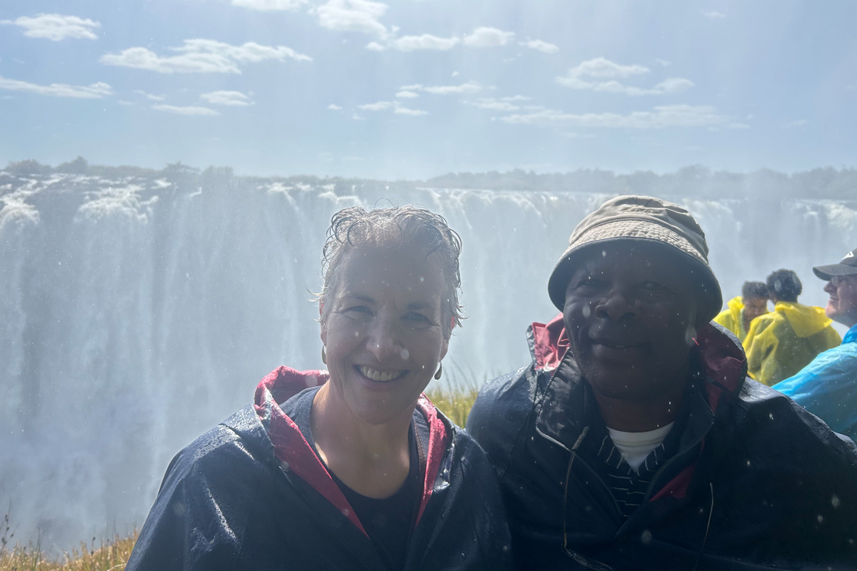 Two smiling tourists in rain jackets standing together at a waterfall viewpoint, mist from the massive falls visible in background along with other visitors in colorful gear.