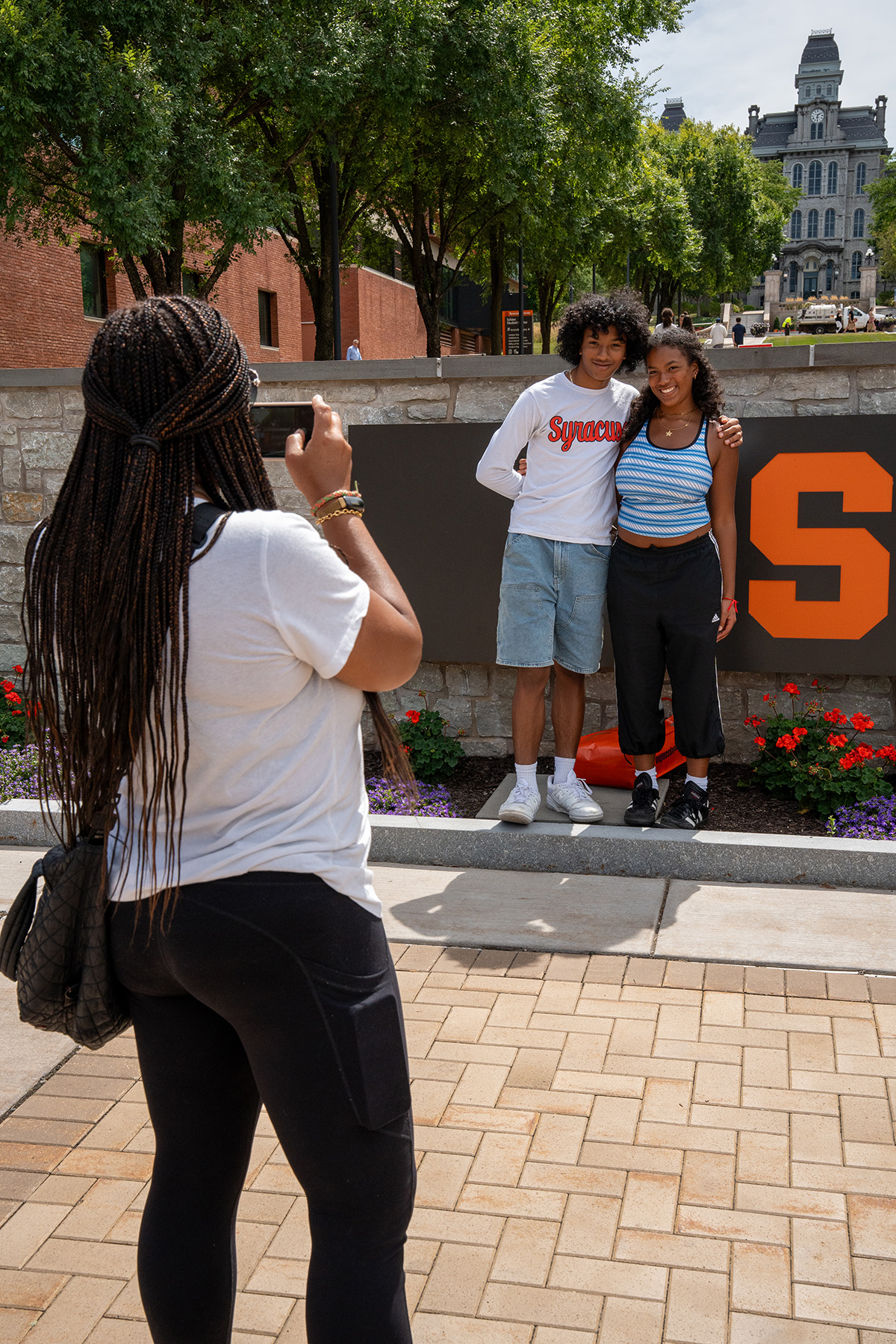 an individual takes a photo of two people in front of the Syracuse University logo sign in front of the walkway leading to the Hall of Languages on the Syracuse University campus. The person posing on the left is wearing a white Syracuse shirt and jean shorts and the person on the right is wearing a blue and white striped tank top and black pants.