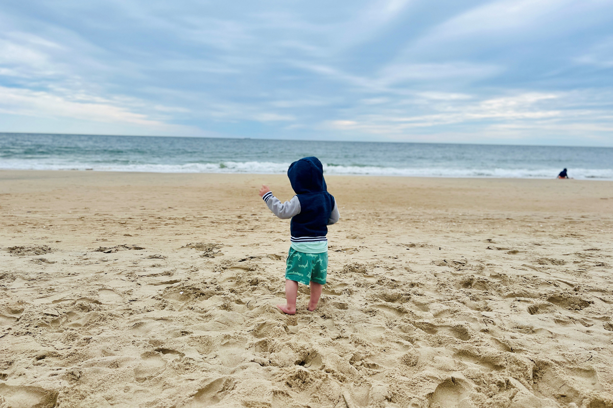 A toddler in a navy hoodie and teal shorts stands on a sandy beach, pointing toward the ocean under a cloudy blue sky.