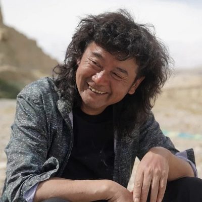 Smiling man with curly dark hair wearing a gray speckled sweater, photographed outdoors with a desert landscape in the background.