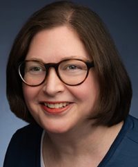 A woman with glasses smiles for a headshot in front of a grey backdrop.