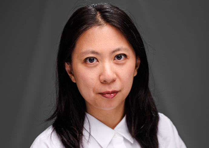 Professional headshot of a person with long dark hair wearing a white collared shirt against a gray background.