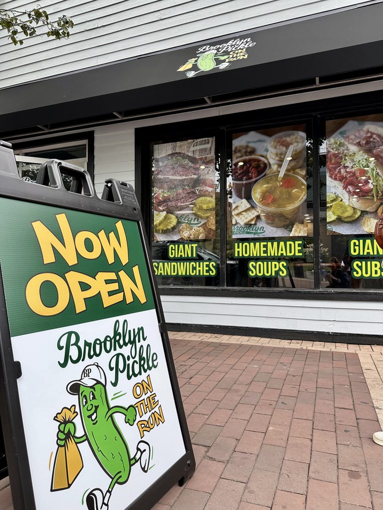 Exterior of Brooklyn Pickle On The Run restaurant with a black awning and sandwich board sign reading “NOW OPEN”; windows display food images and text: “GIANT SANDWICHES,” “HOMEMADE SOUPS,” and “GIANT SUBS.”