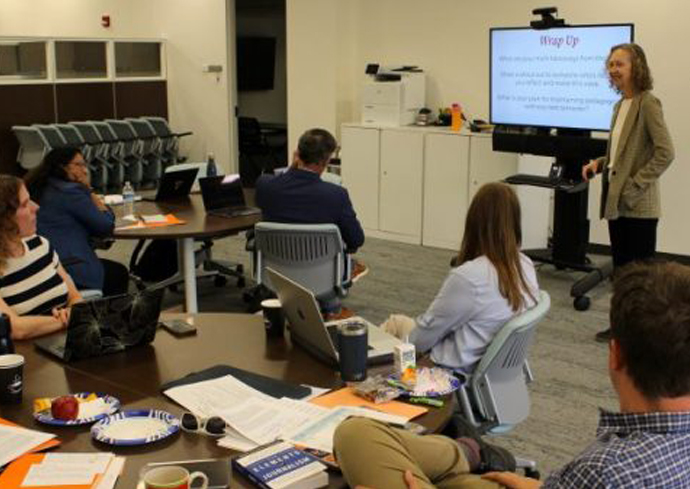 A woman in a beige cardigan presents to a small group seated around a conference table in a modern office space, with a large wall-mounted screen displaying presentation content behind her.RetryClaude can make mistakes. Please double-check responses.