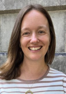 Person with shoulder-length brown hair wearing a striped floral shirt stands in front of a concrete wall. 