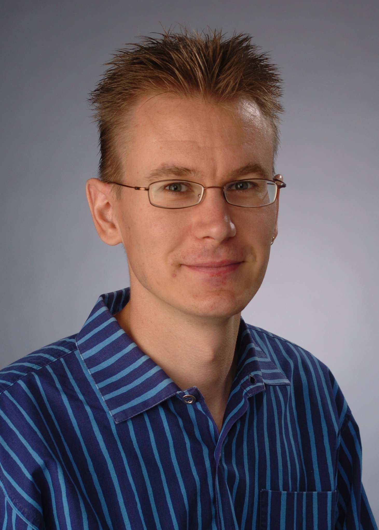 Professional headshot of a man with short spiky blonde hair wearing wire-rimmed glasses and a blue striped button-down shirt against a gray background.