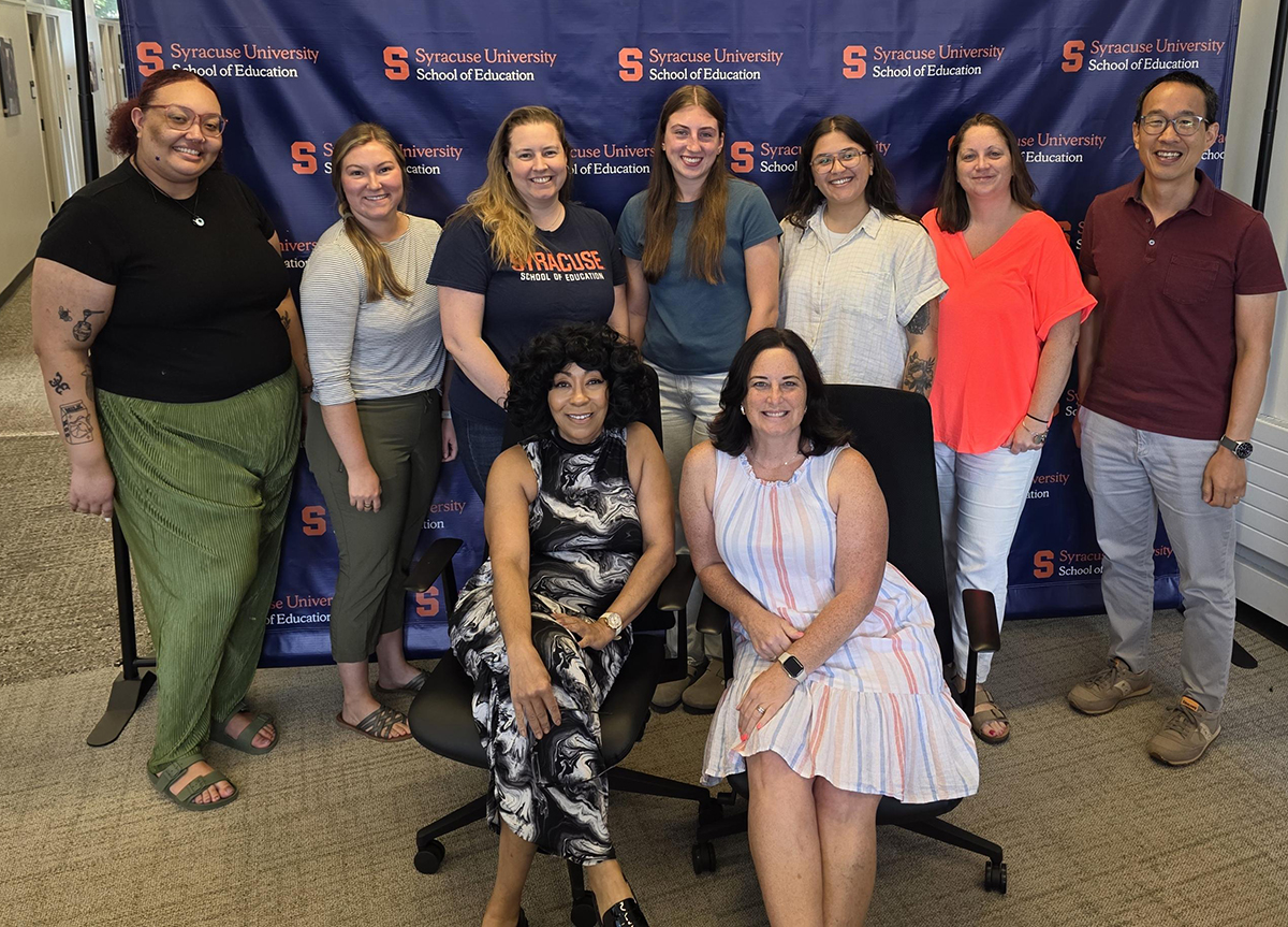 Group photo of nine people in front of a blue 'Syracuse University School of Education' backdrop, with seven standing and two seated.