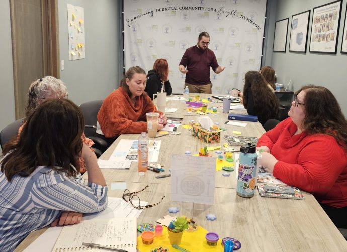 Group of people seated around a conference table with notebooks, drinks, toys, and craft supplies, participating in a workshop led by a presenter. A banner in the background reads 'Empowering Our Rural Community For a Brighter Tomorrow' with 'Family Tree' logos.