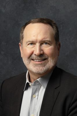 man with short hair and beard wearing a dark suit jacket over a light checkered shirt, posed against a textured dark gray background