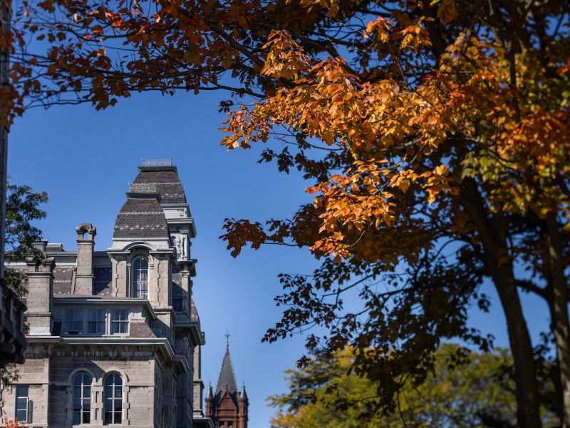 Historic stone building with steep roof and ornate details framed by orange autumn trees under a clear blue sky, with a spired building in the background.