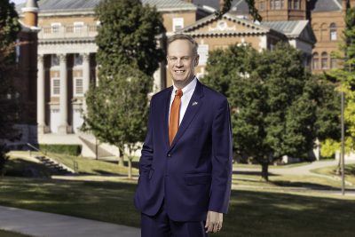 Person in navy suit and orange tie standing on grass in front of large historic building with columns and trees.