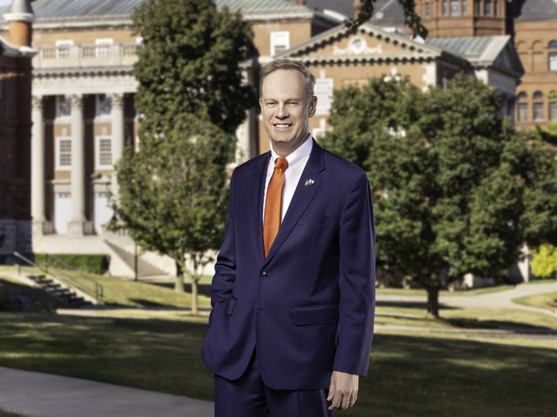 Person in navy suit and orange tie standing on grass in front of large historic building with columns and trees.