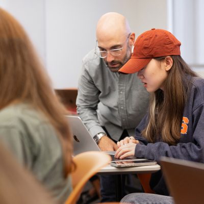 Instructor assisting student wearing orange cap with laptop work in classroom.