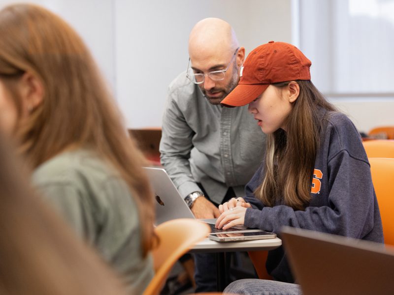 Instructor assisting student wearing orange cap with laptop work in classroom.