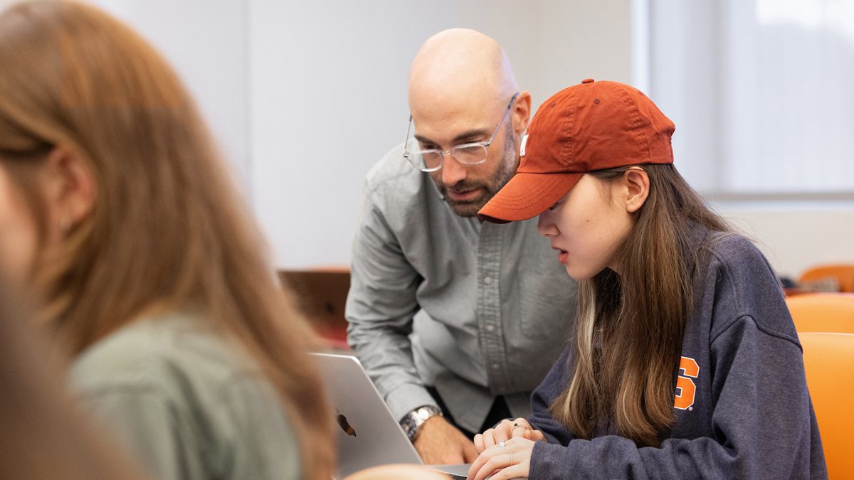 Instructor assisting student wearing orange cap with laptop work in classroom.