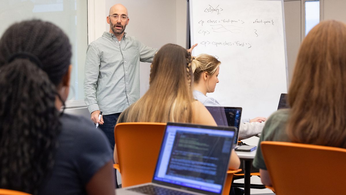 Instructor teaching programming class to students working on laptops with whiteboard code examples.