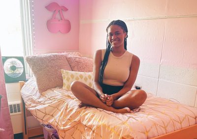 A student sits cross-legged on a bed with golden bedding in a pink-themed bedroom, smiling at the camera. The room features pink walls with cherry decorations and a small fan by the window.