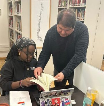 Two people in a library. One is seated and holding a book, the other is standing and helping the other person find information by leafing through the pages. 