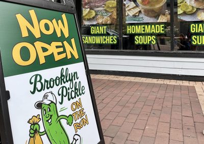 Sidewalk sign outside a restaurant reads “NOW OPEN Brooklyn Pickle ON THE RUN” with a cartoon pickle holding a sandwich bag; background windows display “GIANT SANDWICHES” and “HOMEMADE SOUPS.”