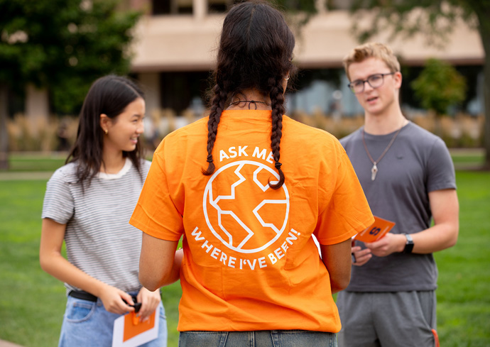 Three people chat with one another outdoors; one wears an orange shirt with 'ASK ME WHERE I'VE BEEN!' and a globe design, facing two others holding pamphlets.