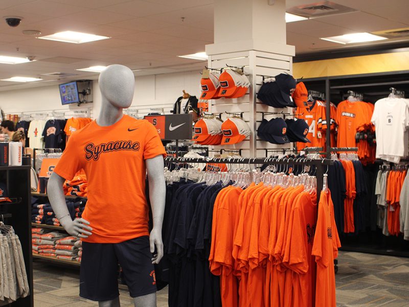 Mannequin wearing Syracuse University orange t-shirt and navy shorts in a merchandise store filled with branded apparel, including t-shirts, hats and jerseys in school colors, with a visible Nike logo on a shelf.