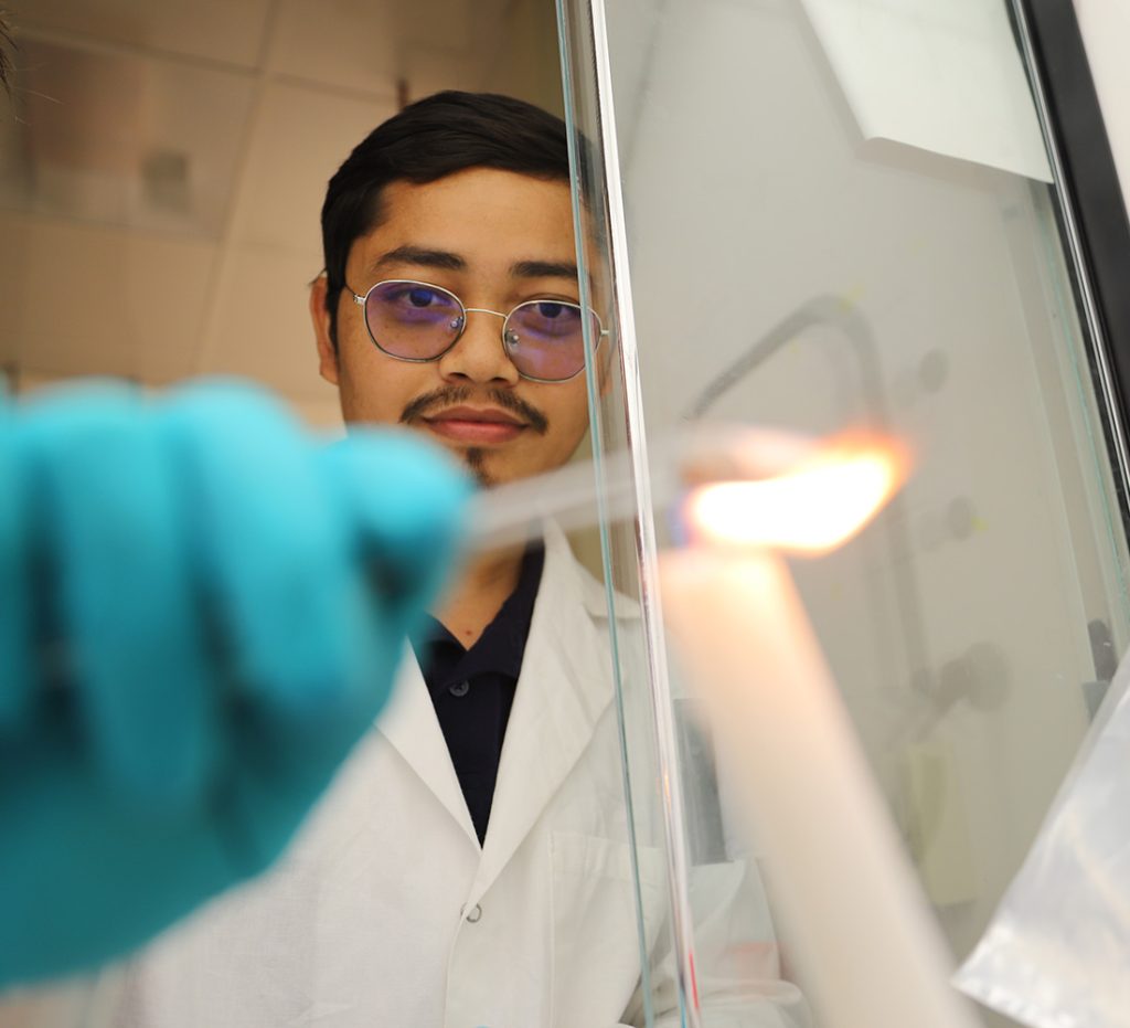 A person in a lab coat and safety glasses observes a lit candle in a laboratory.