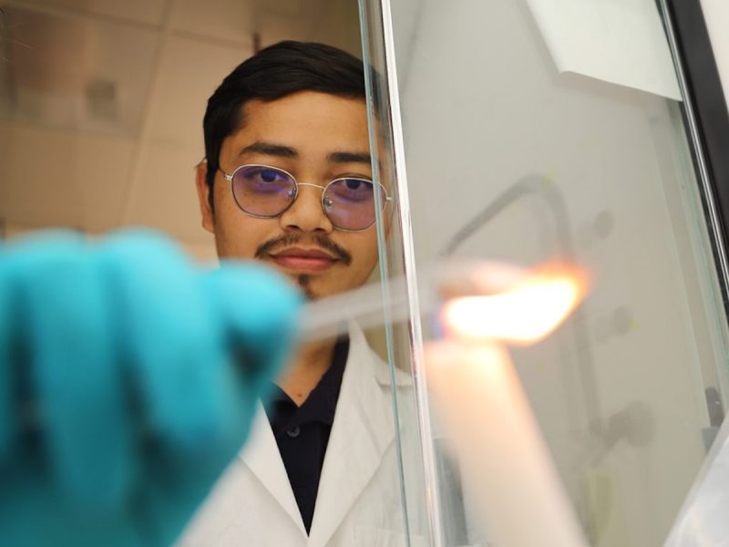 Person in lab coat, glasses and gloves holds a burning candle in a laboratory.